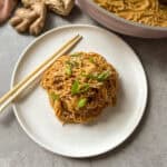 A plate of noodles garnished with green onions and sesame seeds sits next to chopsticks. A bundle of fresh ginger is in the background, along with a pan of additional noodles. The scene is set on a gray countertop.