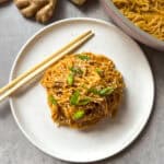 A plate of saucy Peanut Butter Noodles garnished with sesame seeds and green onions, with chopsticks on the side. Fresh ginger and a pan of more noodles are visible in the background on a gray surface.