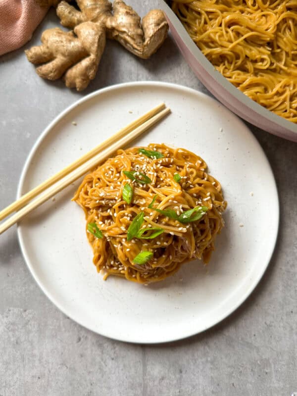 A plate of saucy Peanut Butter Noodles garnished with sesame seeds and green onions, with chopsticks on the side. Fresh ginger and a pan of more noodles are visible in the background on a gray surface.