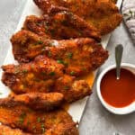 Slices of crispy fried chicken breast are lined up on a white platter, each coated with a glossy, spicy sauce and garnished with chopped herbs. A small white bowl filled with extra sauce and a spoon sits beside the platter.