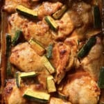 Baked chicken thighs with pieces of zucchini in a rich, golden sauce, displayed in a baking dish. The chicken appears tender with a caramelized finish, and the zucchini is sliced into batons.