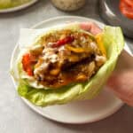 A hand holds a lettuce-wrapped burger filled with grilled patties, sautéed onions, sliced tomatoes, and creamy sauce. The background shows a blurred table setting.