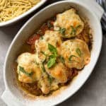 Baked chicken with melted cheese and herbs in a white oval dish. The dish is next to a bowl of cooked spaghetti. A striped cloth is partially visible in the background.