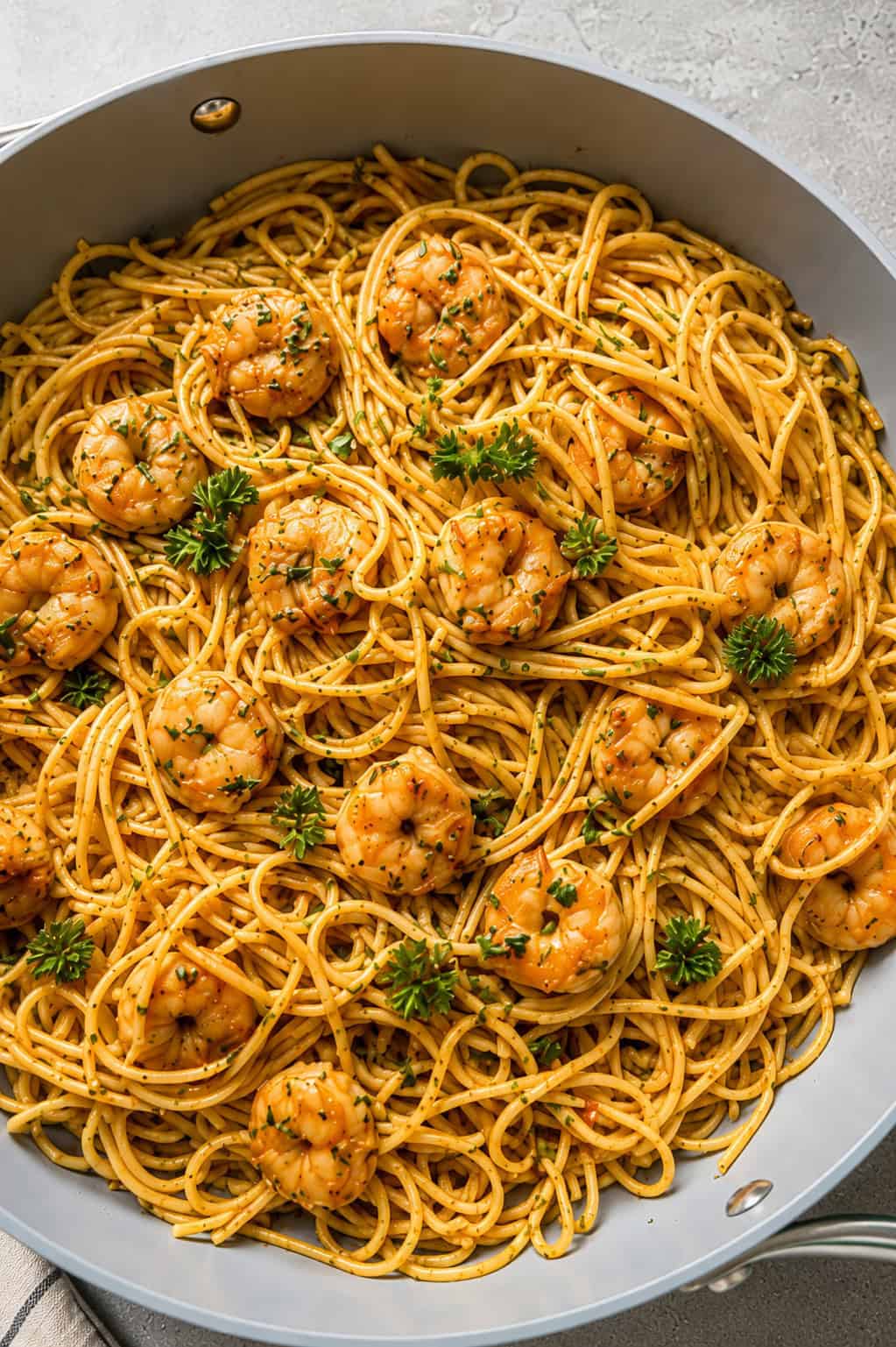 A skillet filled with spaghetti and seasoned shrimp, garnished with fresh parsley, sits on a light-colored surface. The pasta and shrimp are mixed evenly, creating a vibrant and appetizing dish.