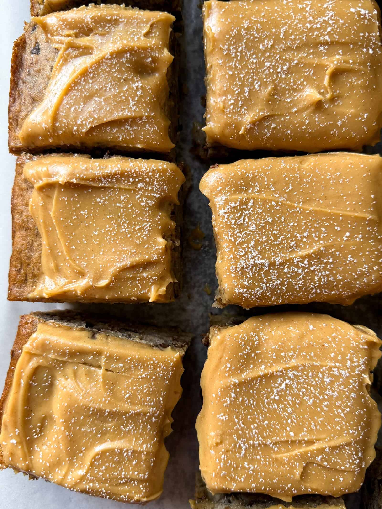 Six rectangular pieces of Banana Chocolate Chip Cake topped with smooth peanut butter frosting and dusted with powdered sugar are arranged on a flat surface.