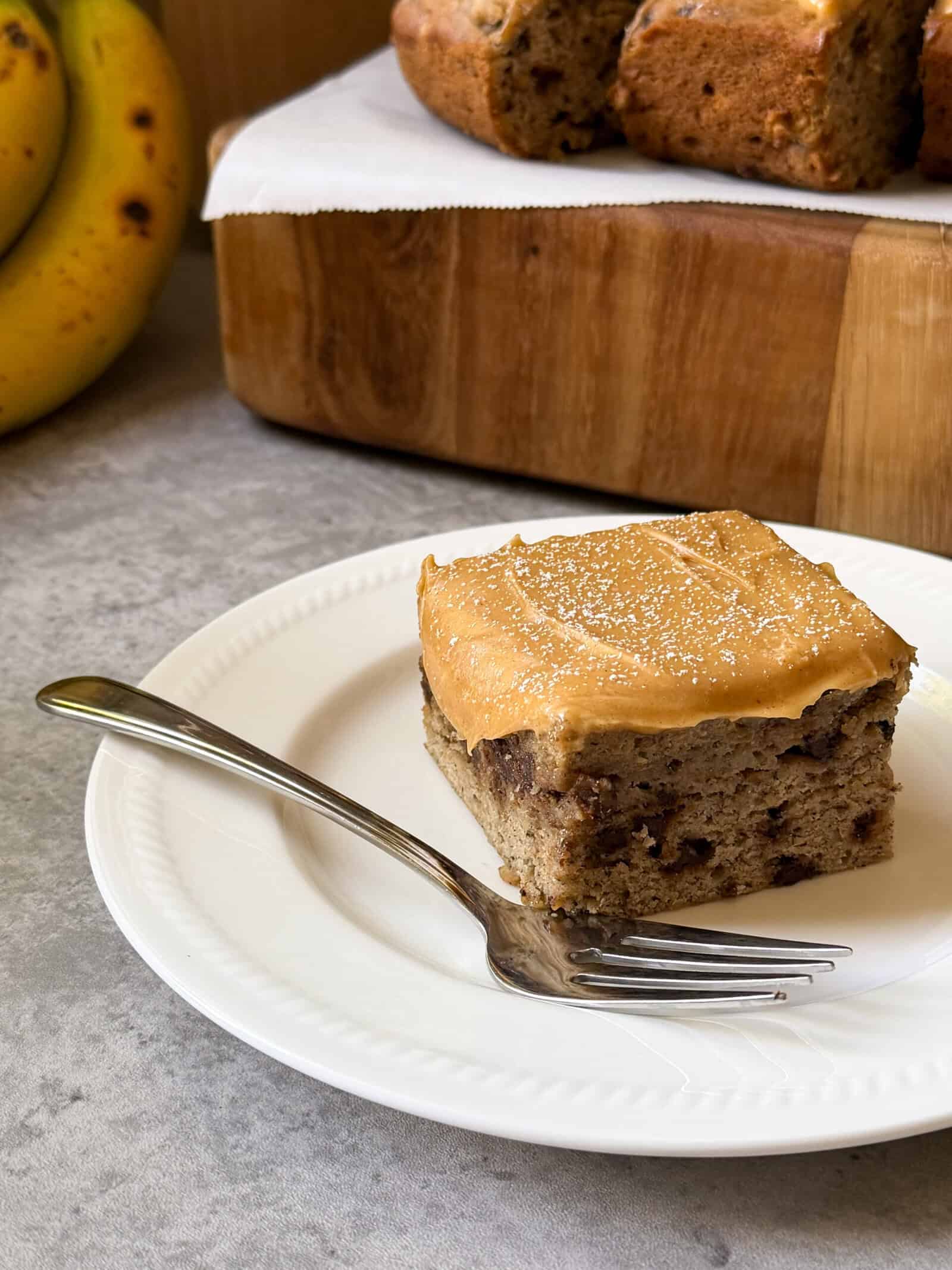 A square slice of Banana Chocolate Chip Cake with tan-colored frosting sits on a white plate with a fork, next to ripe bananas and a wooden board holding more cake in the background.