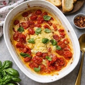 A white baking dish filled with a savory boursin and roasted tomato dip, garnished with fresh basil. Slices of toasted baguette, a bowl of red pepper flakes, a gold spoon, and more basil rest nearby on a gray surface.