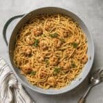 A skillet filled with spaghetti and shrimp in a savory sauce, garnished with fresh herbs, sits on a gray textured surface next to a striped napkin and a pasta serving fork.