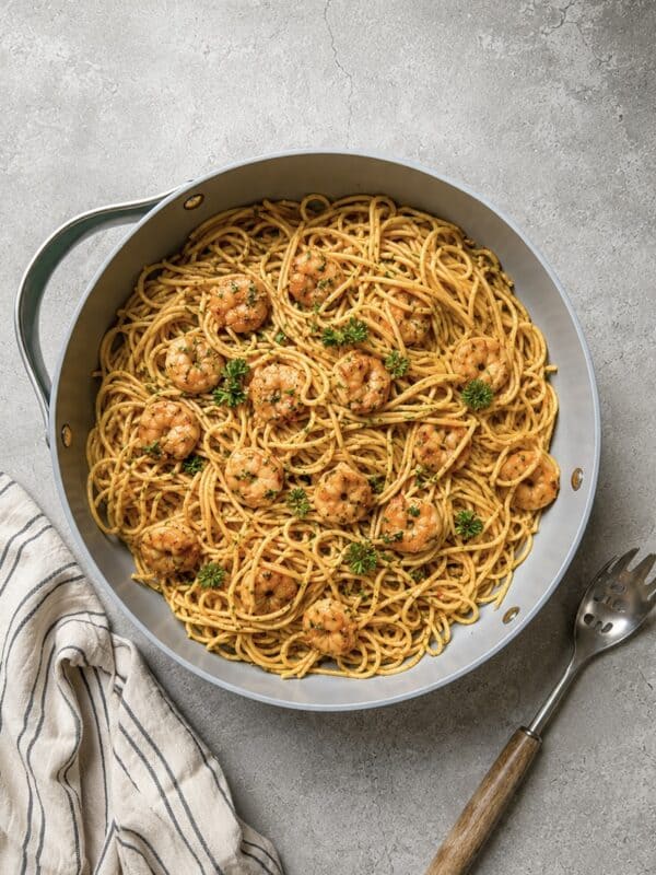A skillet filled with spaghetti and shrimp in a savory sauce, garnished with fresh herbs, sits on a gray textured surface next to a striped napkin and a pasta serving fork.