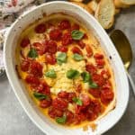 A white oval baking dish filled with roasted cherry tomatoes, a block of melted cheese, and fresh basil leaves. The dish is on a gray surface with slices of baguette and a gold spoon beside it. A white cloth is partially visible on the side.