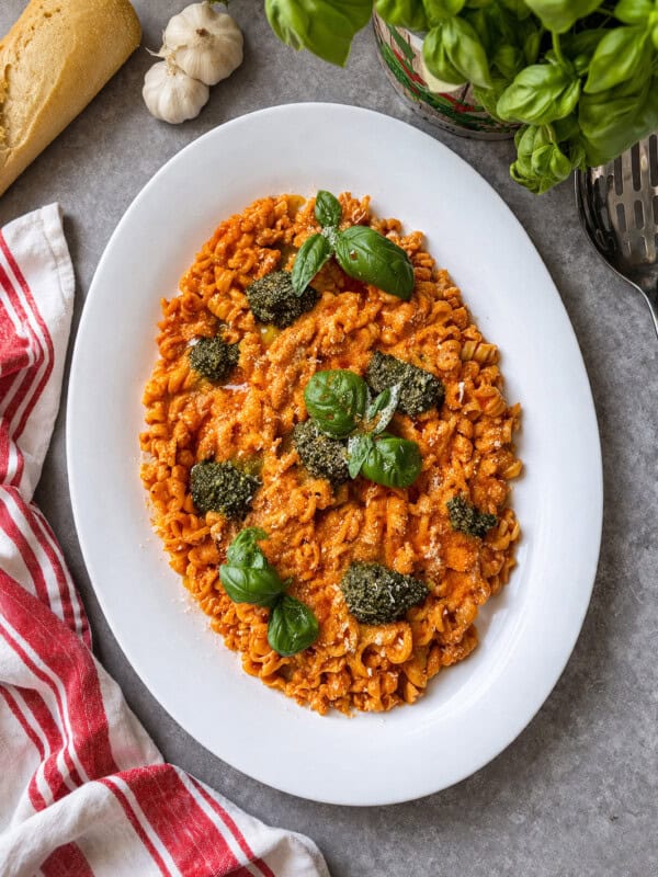A white oval plate of pasta with red sauce, topped with dollops of green pesto and fresh basil leaves, sits on a gray surface near a red-striped towel, garlic, bread, and a basil plant.