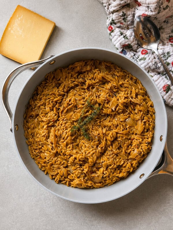 A pan filled with cooked orzo pasta in a rich, brown sauce, garnished with herbs, sits on a light surface next to a wedge of cheese, a floral oven mitt, and a serving spoon.