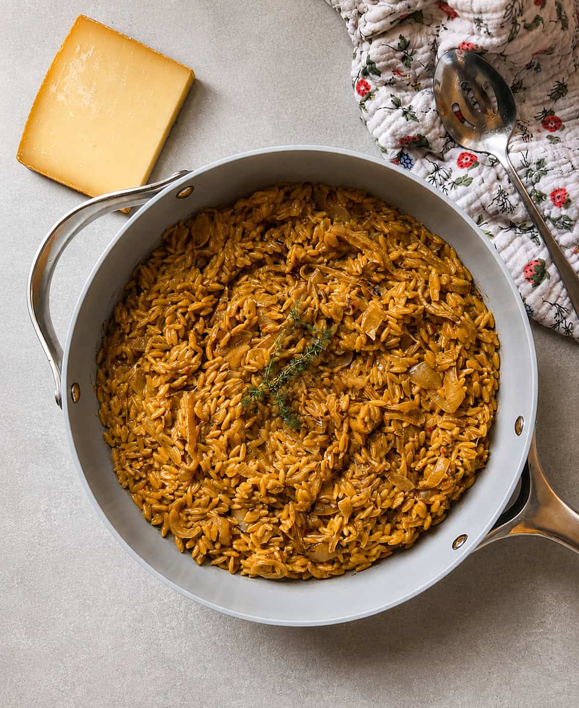 A pan filled with cooked orzo pasta in a rich, brown sauce, garnished with herbs, sits on a light surface next to a wedge of cheese, a floral oven mitt, and a serving spoon.
