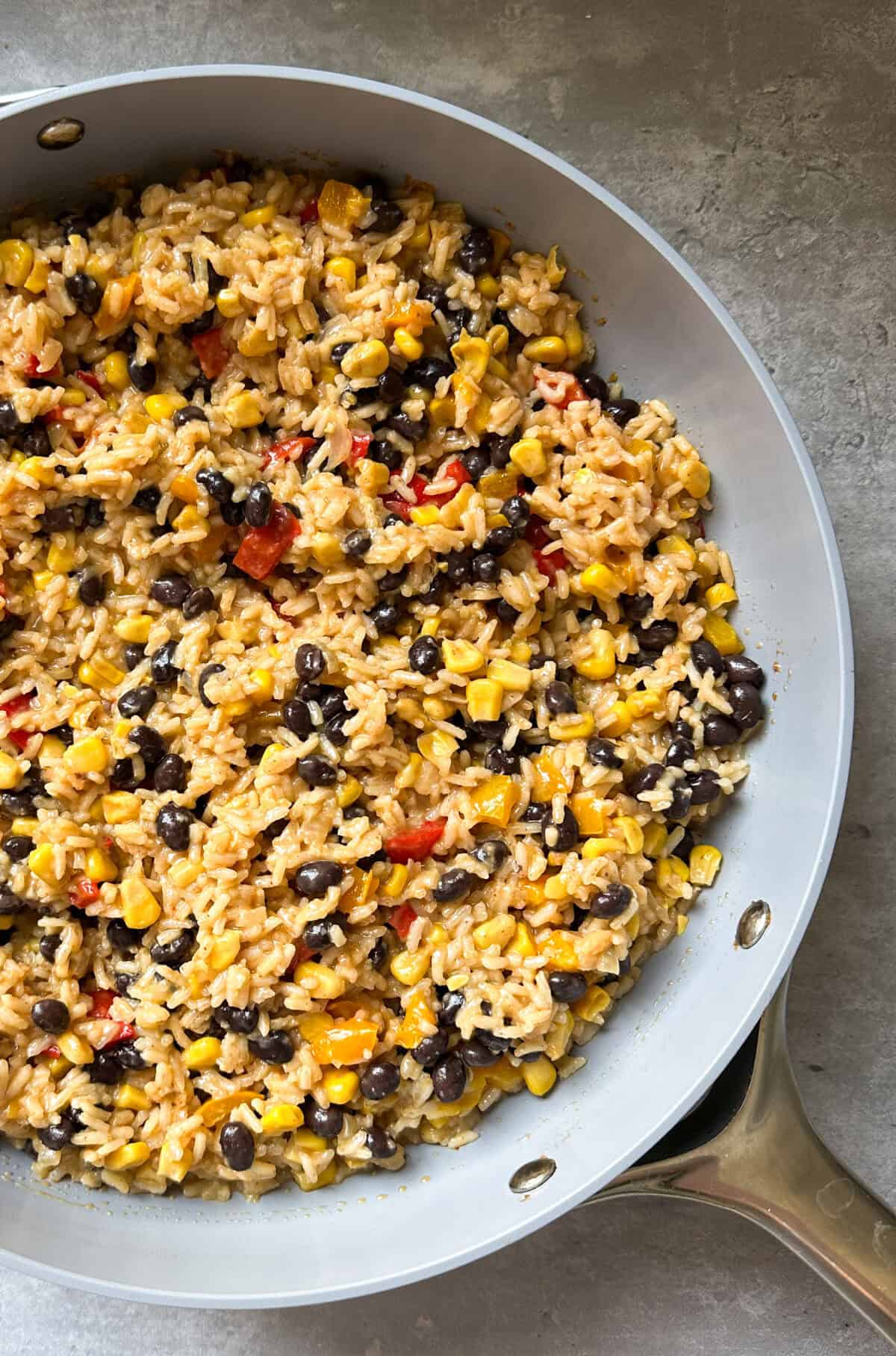 A blackened chicken recipe skillet filled with a colorful mixture of rice, black beans, corn, and small pieces of red and yellow bell pepper on a gray surface.