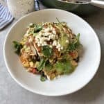 A white plate with sliced blackened chicken breast on a bed of corn, black beans, and rice, topped with green sauce, crumbled cheese, and fresh cilantro—a delicious Blackened Chicken and Rice dish. A pan and a mug are partially visible in the background.