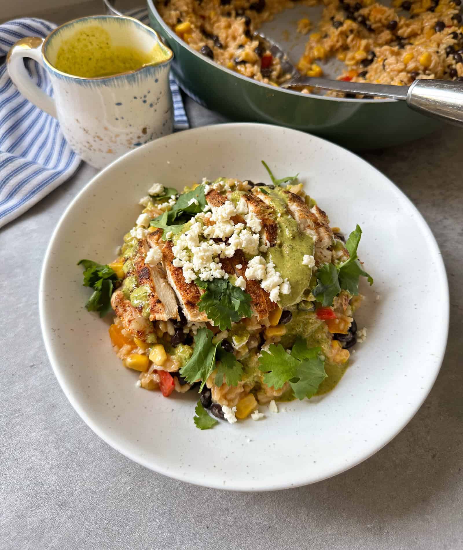 A plate of Mexican-inspired rice casserole topped with grilled chicken, crumbled cheese, fresh cilantro, and green sauce. A skillet with more casserole and a cup of extra sauce are in the background.