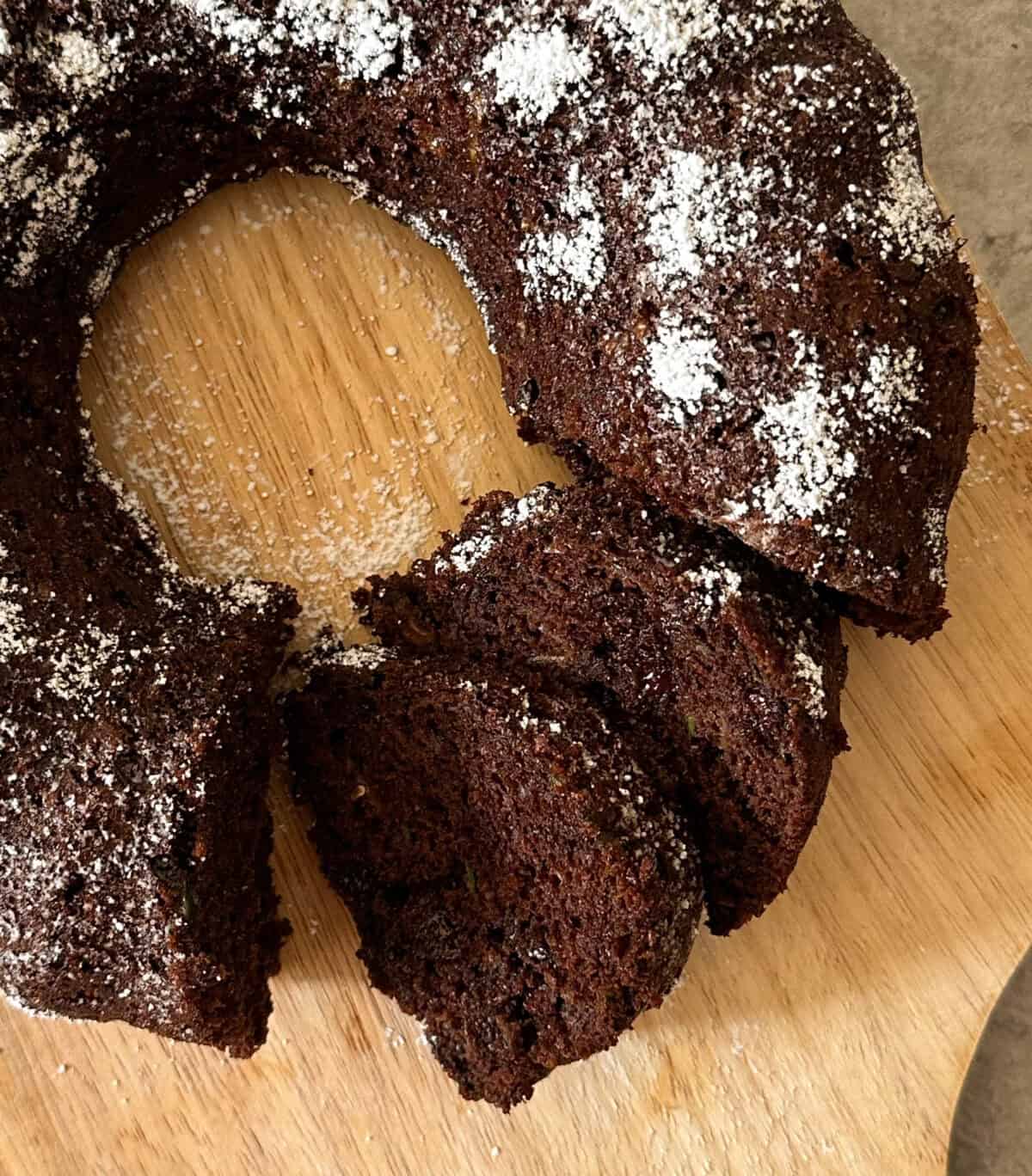 A close-up of a chocolate bundt cake with powdered sugar on top, placed on a wooden board. Several slices have been cut and are lying in front, reminiscent of a rich Chocolate Chip Zucchini Bread.