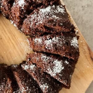 A close-up of sliced Chocolate Zucchini Bread, topped with powdered sugar, arranged on a wooden cutting board.