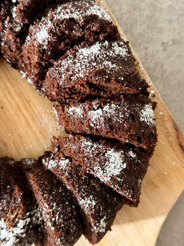 A close-up of sliced Chocolate Zucchini Bread, topped with powdered sugar, arranged on a wooden cutting board.