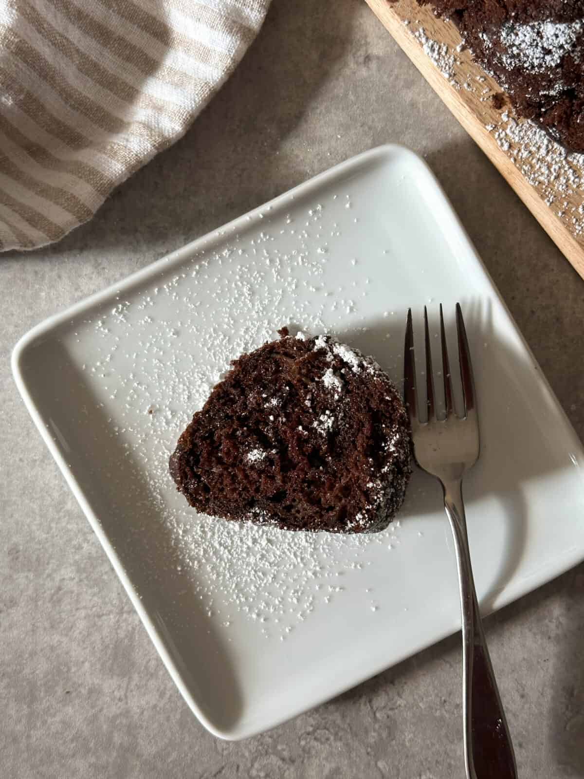 A slice of Chocolate Chip Zucchini Bread, dusted with powdered sugar, sits on a square white plate with a fork beside it. Part of a striped cloth and a wooden board with more bread are visible in the background.