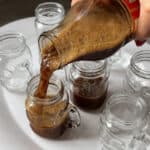 A hand pours a rich, frothy Italian coffee from a bottle into a small glass mug on a tray. Several other empty or partially filled glass mugs are arranged around it, ready to be enjoyed.