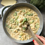 A hand holds a wooden spoon scooping creamy White Cheddar Broccoli Mac and Cheese from a gray pan. A block of cheese and a head of broccoli are visible nearby on the countertop.