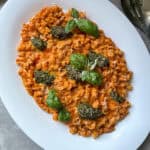 Plate of pasta with red sauce, topped with green herbs and basil leaves, served on a white oval platter. A metal serving spoon is partially visible to the side.