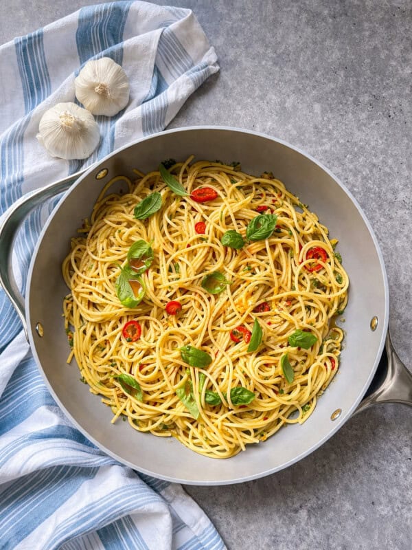 A skillet filled with cooked spaghetti aglio e olio, garnished with fresh basil and sliced red chili, sits on a gray surface next to two garlic bulbs and a blue-and-white striped cloth.
