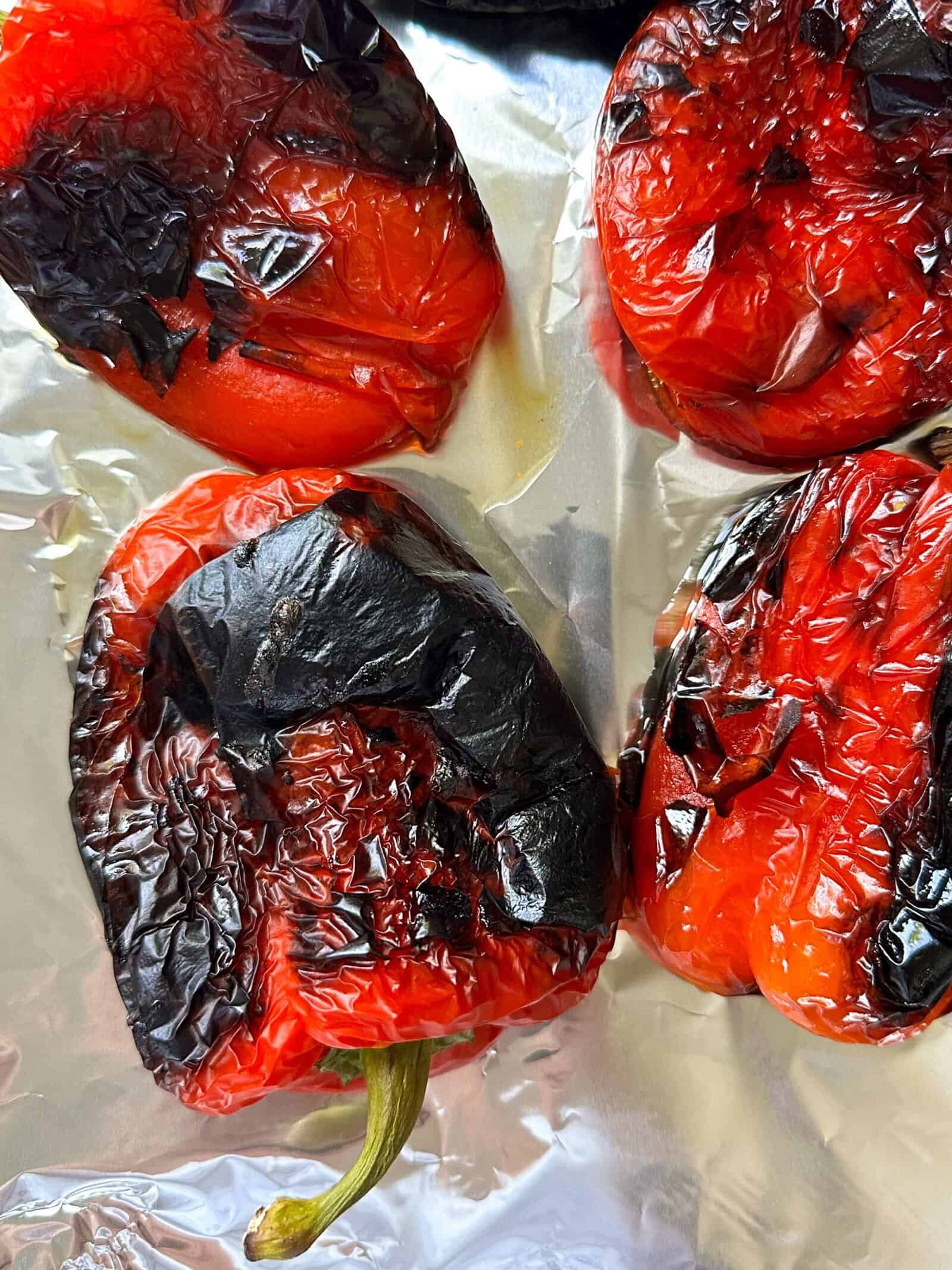 Four roasted red peppers with charred, blistered skins rest on a sheet of aluminum foil. The peppers show blackened spots from roasting, with wrinkled and slightly shiny surfaces—perfect for any roasted peppers recipe.