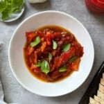 A white plate with roasted red peppers in oil, garnished with fresh basil leaves. Nearby are a head of garlic, extra basil on a small plate, a red container, and slices of bread—perfect for trying your favorite roasted peppers recipe.