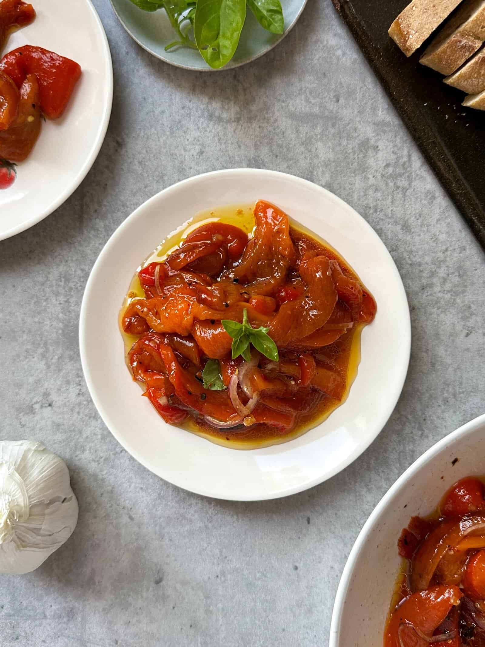 A white plate with marinated roasted red peppers in olive oil, garnished with fresh basil, sits on a gray surface. Nearby are slices of bread, more plates of this roasted peppers recipe, a garlic bulb, and basil leaves.