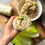 A hand holds a lettuce wrap filled with creamy Jalapeño Popper Chicken salad. Below, a bowl of jalapeno popper chicken salad, a wrap, and crisp romaine lettuce leaves are arranged on a wooden board.