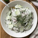 A bowl of Creamy Cucumber Salad with Mayo features crisp cucumbers, red onions, and fresh dill, served on a wooden surface with a spoon, lemon half, and striped cloth for a refreshing touch.
