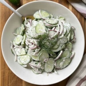 A bowl of Creamy Cucumber Salad with Mayo features crisp cucumbers, red onions, and fresh dill, served on a wooden surface with a spoon, lemon half, and striped cloth for a refreshing touch.