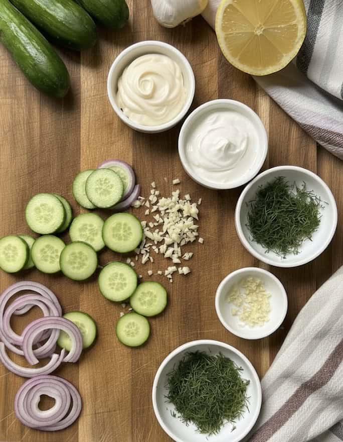 Sliced cucumbers, red onions, minced garlic, fresh dill, mayonnaise, and yogurt are arranged on a wooden cutting board—everything you need to create the perfect Creamy Cucumber Salad Recipe. A lemon half and striped towel add a fresh touch.