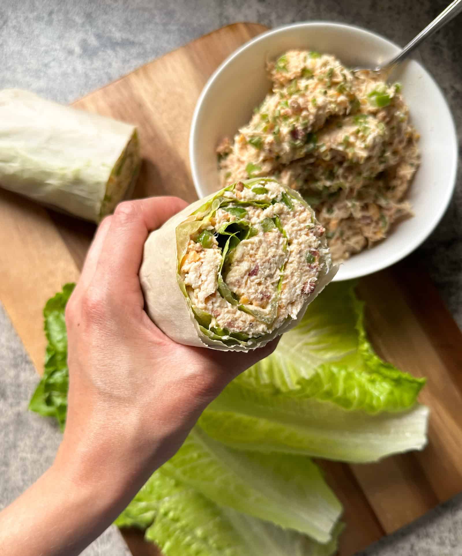 A hand holds a wrap filled with jalapeño popper chicken lettuce wraps. A halved wrap and a white bowl containing more of the zesty chicken mixture are on a wooden cutting board, surrounded by loose lettuce leaves.