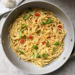 A pan filled with spaghetti aglio olio e peperoncino, mixed with sliced red chili peppers and garnished with fresh basil. The pasta is lightly coated with oil, a garlic bulb visible in the corner. All set against a grey countertop background.