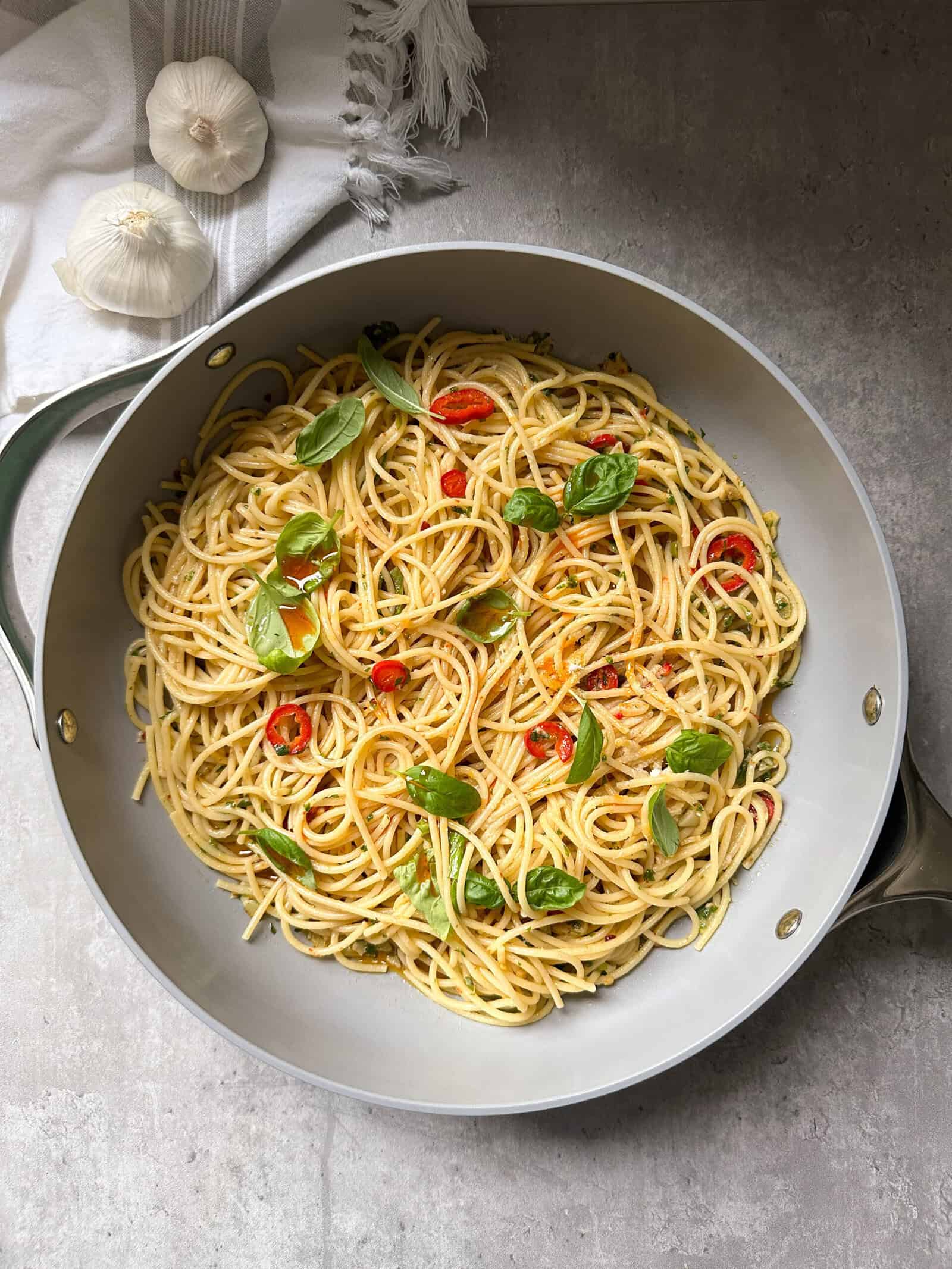 A skillet filled with spaghetti aglio olio e peperoncino, garnished with fresh basil and sliced red chili peppers, sits on a gray surface. A white cloth and two garlic bulbs are visible in the upper left corner.