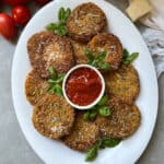 A white platter with breaded, golden-fried red tomatoes garnished with basil and grated cheese, arranged around a small bowl of marinara sauce. Fresh garden tomatoes and a wedge of cheese are nearby.