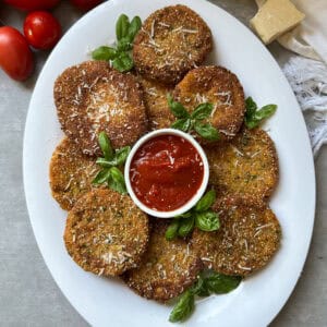 A white platter with breaded, golden-fried red tomatoes garnished with basil and grated cheese, arranged around a small bowl of marinara sauce. Fresh garden tomatoes and a wedge of cheese are nearby.