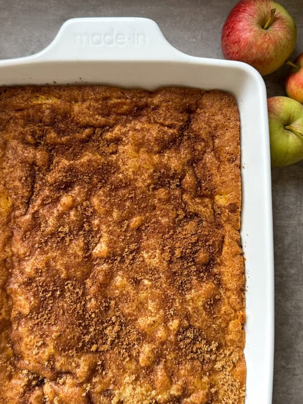 A golden-brown Apple Cake topped with cinnamon sugar in a white made-in dish sits next to three apples on a gray surface.