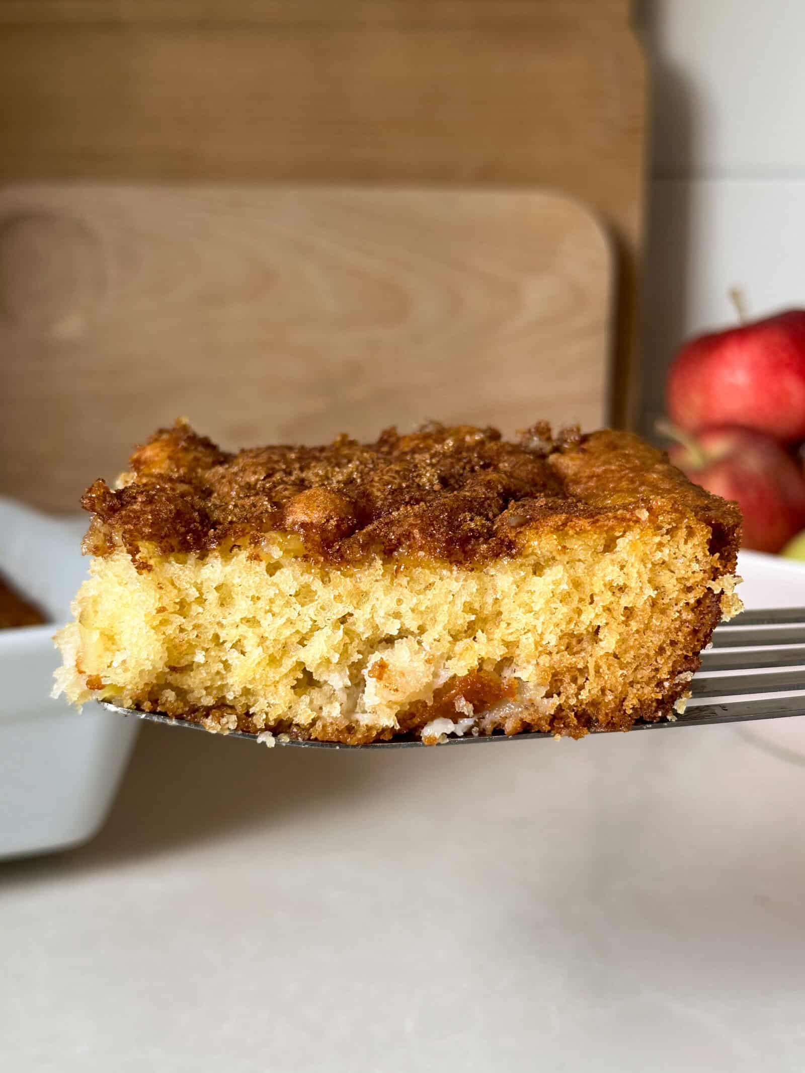 A close-up of a slice of moist apple cake with a crumbly brown topping, held on a spatula. In the background, red apples, a wooden cutting board, and a white baking dish suggest an easy apple cake recipe in progress.