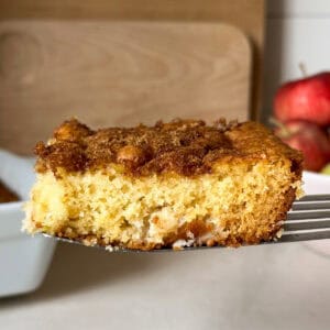 A close-up of a slice of moist apple cake with a cinnamon sugar topping, held up on a spatula. In the background, apples and a wooden cutting board hint at the fresh ingredients in this easy apple cake.