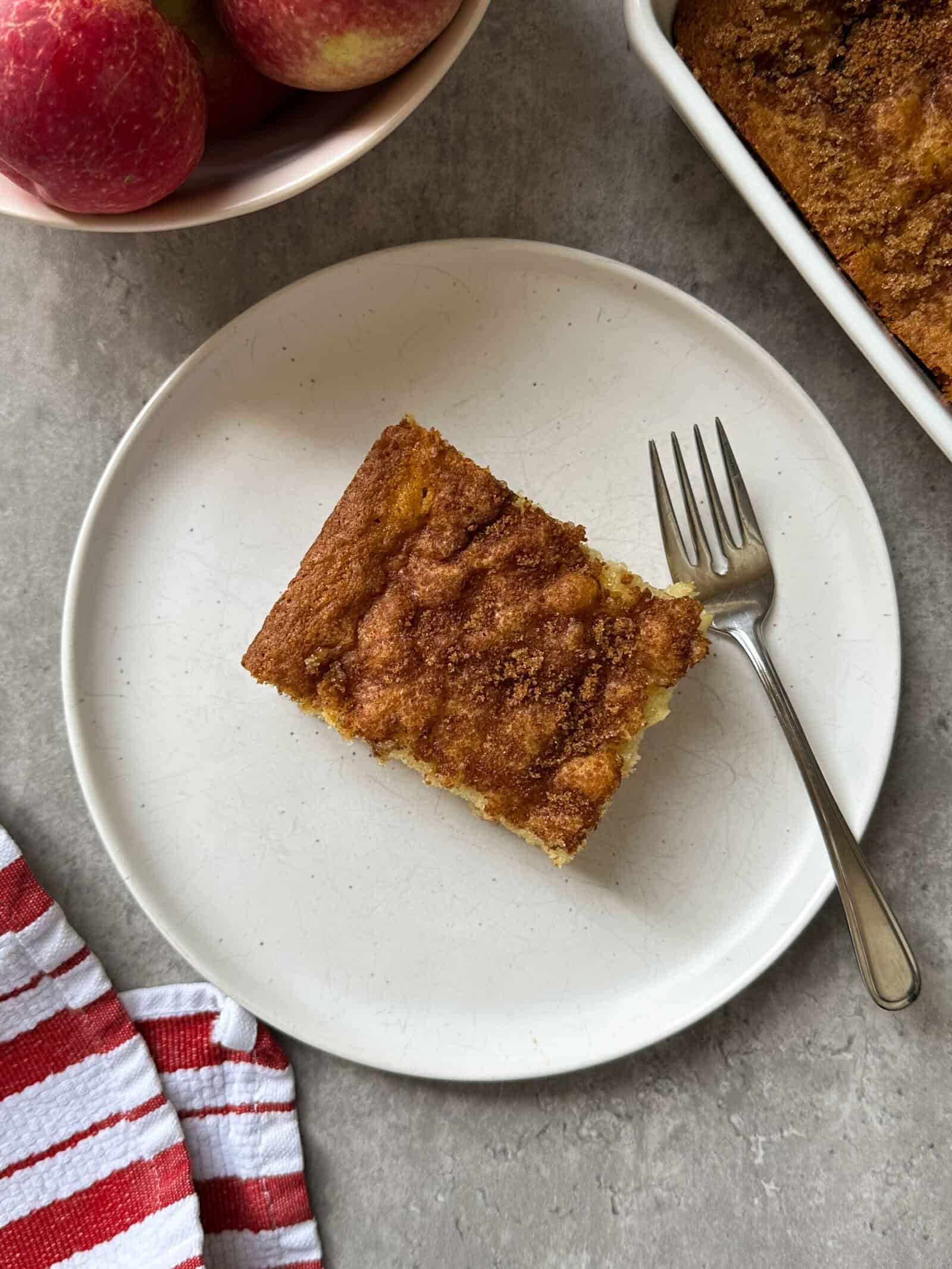 A slice of cinnamon-topped Easy Apple Cake on a white plate with a fork, beside a red-and-white striped cloth, a bowl of apples, and part of a baking dish, all set on a gray surface.