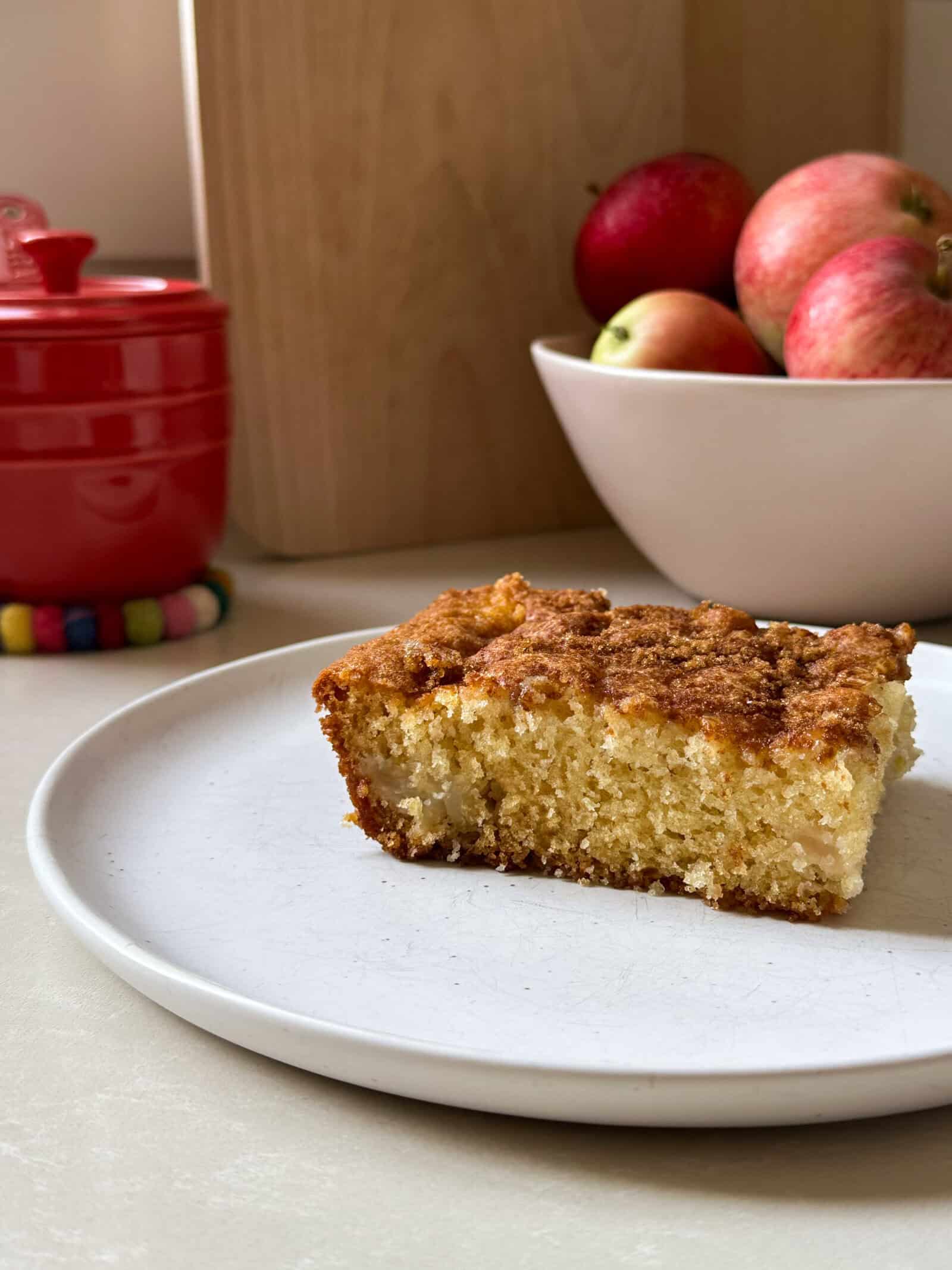 A slice of moist apple cake sits on a white plate. In the background, a bowl of red apples and a red container rest on the countertop.