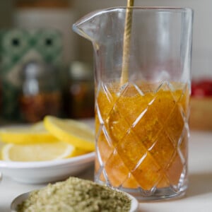 A glass pitcher with a golden stirring spoon holds a yellow-orange barone house dressing mixture. In the foreground, there are small plates with dried herbs and lemon slices, set on a kitchen counter.