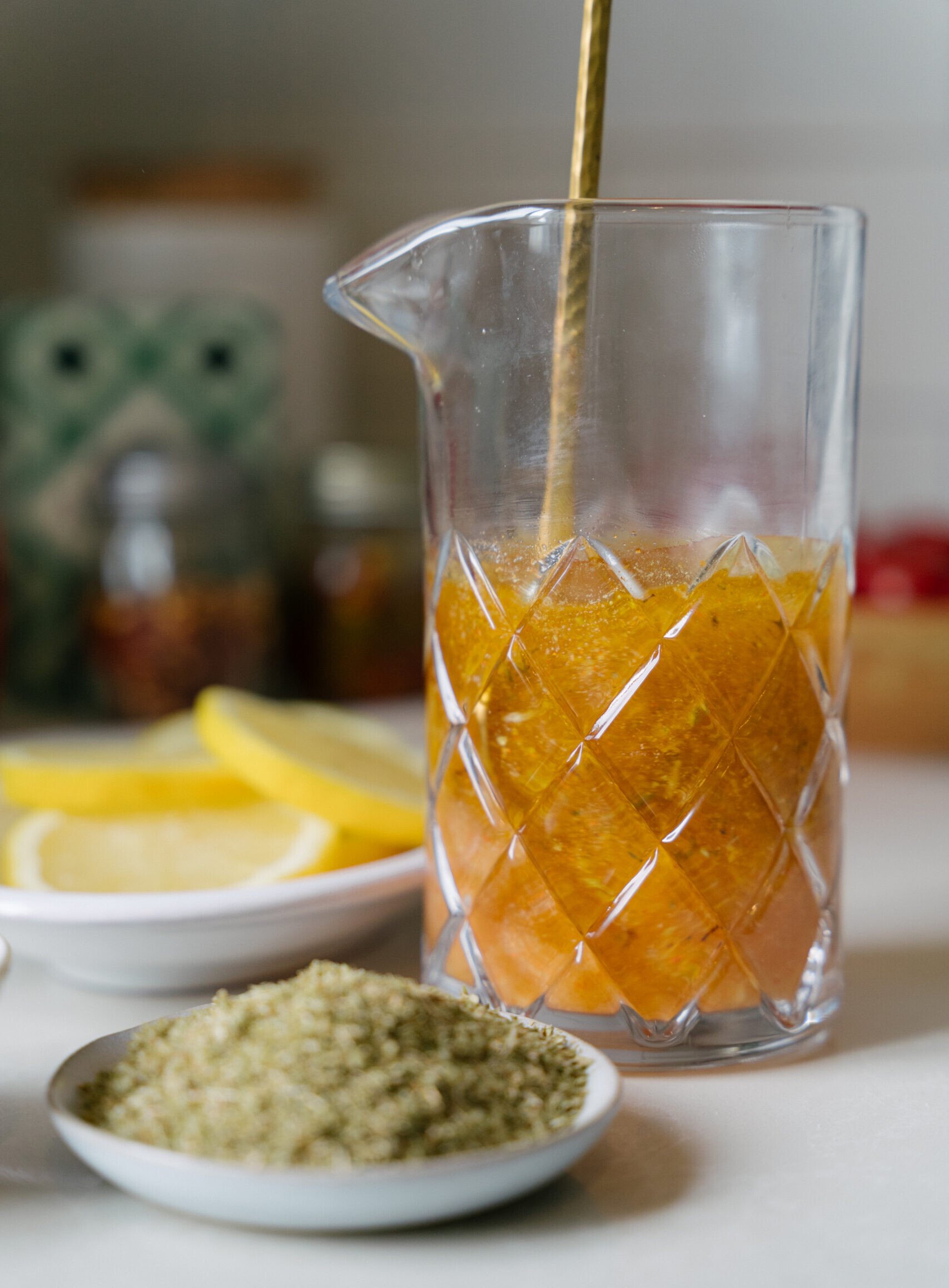 A glass pitcher with a golden stirring spoon holds a yellow-orange barone house dressing mixture. In the foreground, there are small plates with dried herbs and lemon slices, set on a kitchen counter.