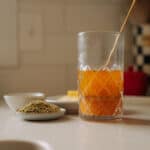 A glass of orange lemon vinaigrette with a stir stick sits on a kitchen counter near small plates of dried herbs and lemon slices, with a blurred kitchen background.