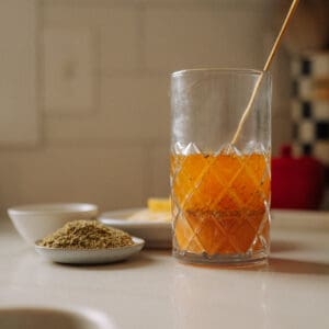 A glass of orange lemon vinaigrette with a stir stick sits on a kitchen counter near small plates of dried herbs and lemon slices, with a blurred kitchen background.