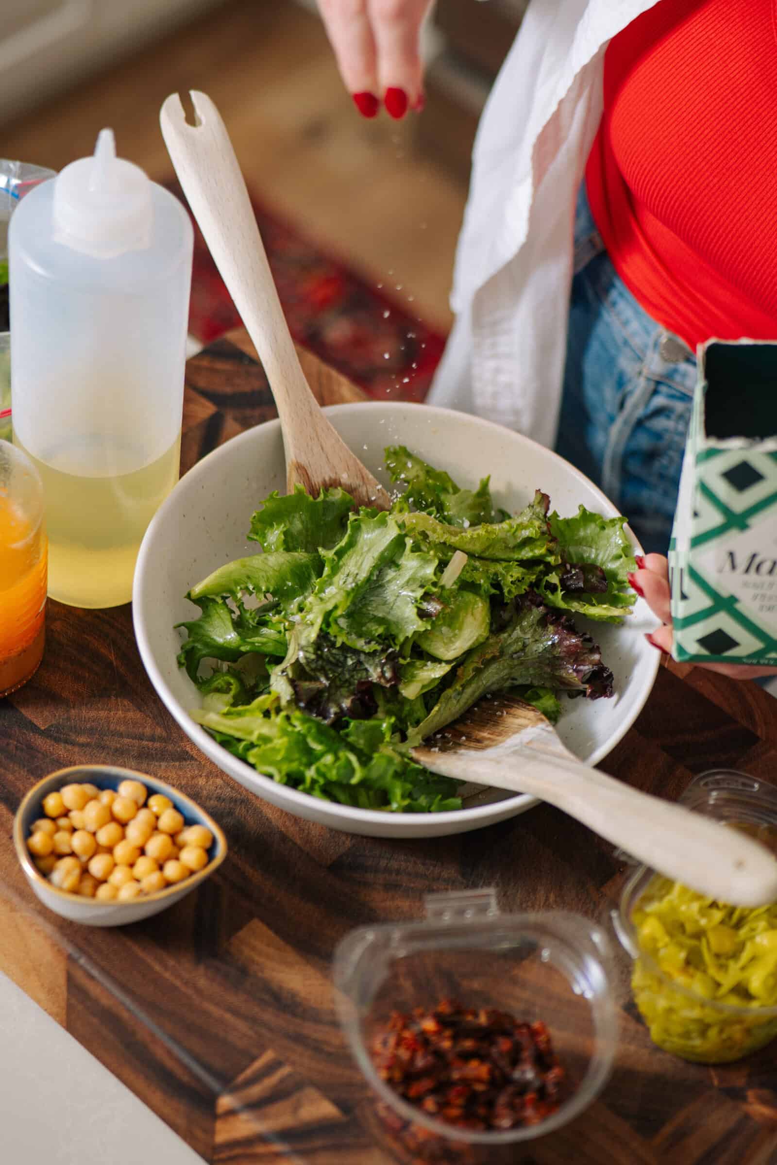 A person prepares a fresh green salad in a white bowl, mixing with wooden utensils. Nearby on the wooden counter are various salad toppings like chickpeas, sun-dried tomatoes, and a tangy lemon vinaigrette.